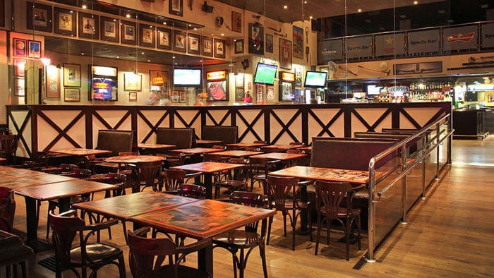 The inside of a bar and pub with wooden chairs or a high-back bench around each of the tables, four televisions hung from the ceiling, and pictures hanging on the wall.
