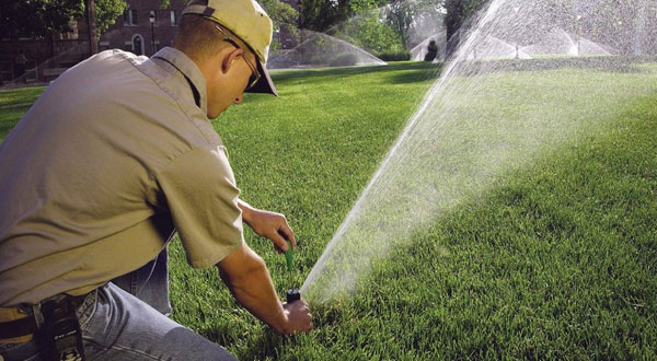 Man with a baseball cap and glasses holding a screw driver to a valve of a lawn sprinkler. The sprinkler is on and spraying away from the man.