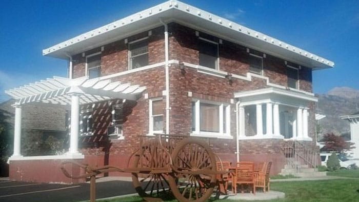 A red brick building with white trim, white pillars, a white open pergola, 10 windows, and stairs leading up to the door with a green lawn and iron wagon wheels and a wooden table with 4 chairs on a patio.