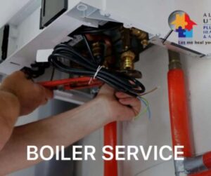 Two hands working on boiler pipes. One hand is holding a red wrench and the other is pushing below the wrench to place it on a valve. A black electrical cord is wrapped by a white cable tie to keep it out of the way.