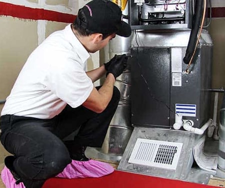 A man in a black baseball cap, white shirt, and black pants checking a furnace.