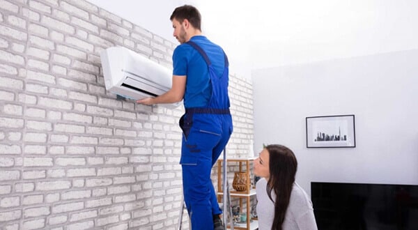 A man in a blue uniform placing an air conditioning wall unit on a brick wall while a lady watches.