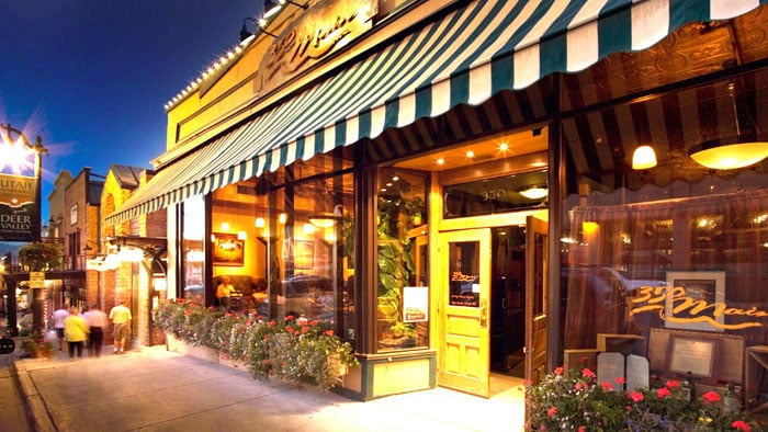 The outside of a bar and grill at night with decorative lights, 350 Main in gold lettering on one of the three large windows, double doors with one open, and a green and white striped awning.