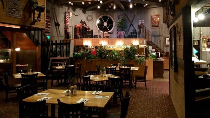 The inside of a restaurant that has decorative lights hanging from the ceiling, plants, and six tables with four wooden chairs each.